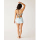 A woman faces away, wearing Carve's Hoku Swim Skirt in light blue with a built-in bikini bottom and a floral halter bikini top; one arm is raised, the other at her side, against a plain background.