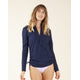 A woman stands against a plain background wearing the Carve Cruz Rashguard in navy with zip front and light purple bikini bottoms.