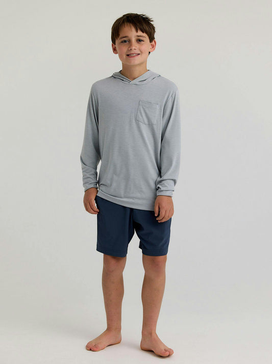 A boy stands barefoot against a plain background, wearing a light gray Free Fly Youth Bamboo Shade Hoody with UPF 50+ sun protection and dark blue shorts.