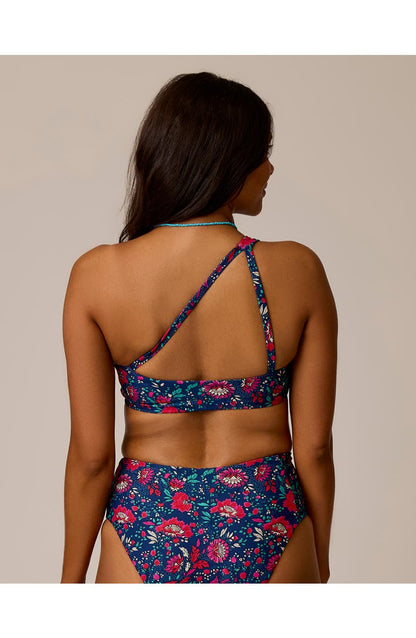 A woman faces away in the Carve Bondi Compression Top, a blue floral, one-shoulder bikini made from recycled compression fabric, photographed against a plain background.