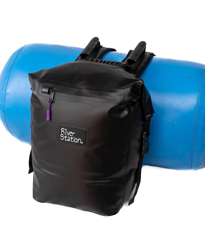 A black waterproof River Station Gear Dry Thwart Bag - V2 backpack with a purple zipper pull in front of a blue barrel.