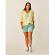 A woman stands smiling in a light yellow sweater, Carve Oahu 6" Twill Short in light green, layered necklaces, and white sneakers against a plain background.