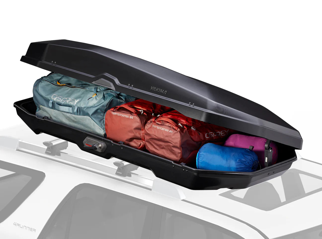 A Yakima CBX 16 roof cargo box is installed on the car, holding four packed bags—among them a teal one and two red ones—with its lid open.