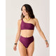 A woman stands smiling against a plain light background, wearing Carve’s maroon Lani Pocket Compression Bottom bikini with a subtle pattern, paired with a gold necklace, bracelets, and watch.
