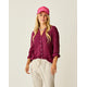 A woman stands against a plain background wearing the Carve Dylan Gauze Tunic in magenta, paired with light pants, a pink cap, and a shell necklace.
