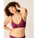 A woman with curly hair poses in the Carve Elba Top by Carve, a maroon seamless bralette bikini, styled with layered shell necklaces, against a plain background; one hand touches her hair while the other rests on her hip.