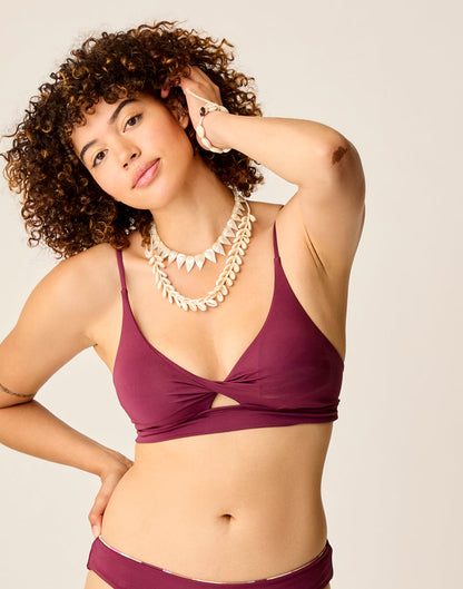 A woman with curly hair poses in the Carve Elba Top by Carve, a maroon seamless bralette bikini, styled with layered shell necklaces, against a plain background; one hand touches her hair while the other rests on her hip.