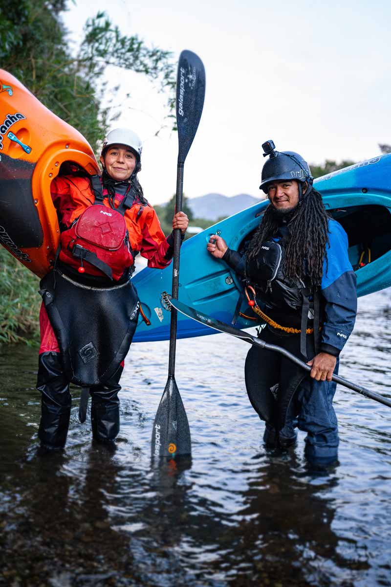 Two people in kayaking gear stand in shallow water, each holding a paddle and carrying a kayak, showing off their 4CRS Rental Drysuits with trees and sky in the background.