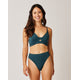 A woman stands smiling against a light background, wearing the Carve Elba Top by Carve—a dark teal, seamless bralette bikini top paired with matching high-waisted bottoms.