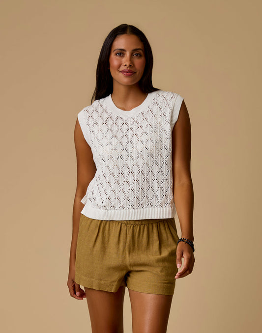 A woman models the Carve Fox Sweater, a sleeveless white knit top, paired with tan shorts against a plain beige background.