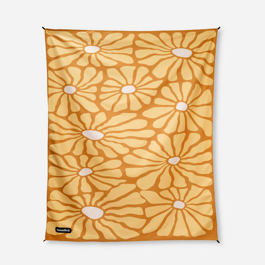 The Nomadix Festival Blanket is a rectangular throw with a yellow and orange floral pattern featuring large stylized flowers with white centers on a plain background.