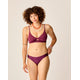 Wearing Carve's Sanitas 2.0 Compression Bottom in burgundy, a person with curly hair accessorizes with jewelry and stands against a plain backdrop.