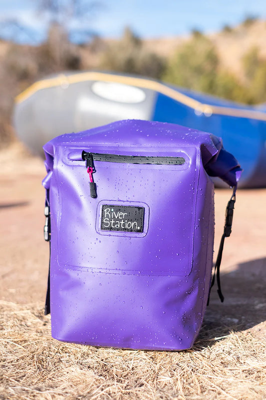 A purple Dry Thwart Bag - V2 with the logo "River Station Gear" on the front, placed on the ground with a canoe visible in the background.