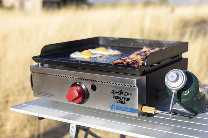 A Camp Chef VersaTop 14 Flat Top Camp Stove with eggs and bacon cooking on its non-stick True Seasoned surface.