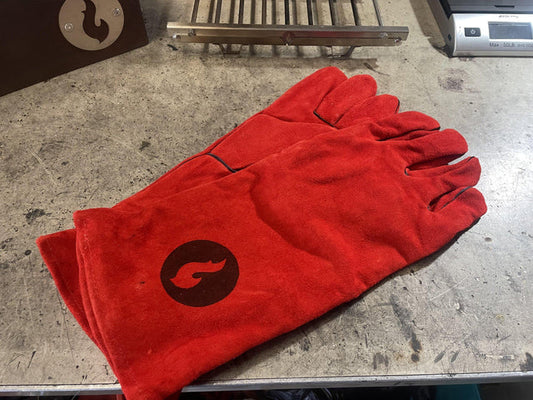LavaBox Fireproof Gloves with a flame logo offer outstanding heat protection for welding tasks; this red heavy-duty leather pair rests on a workbench beside metal tools and equipment.