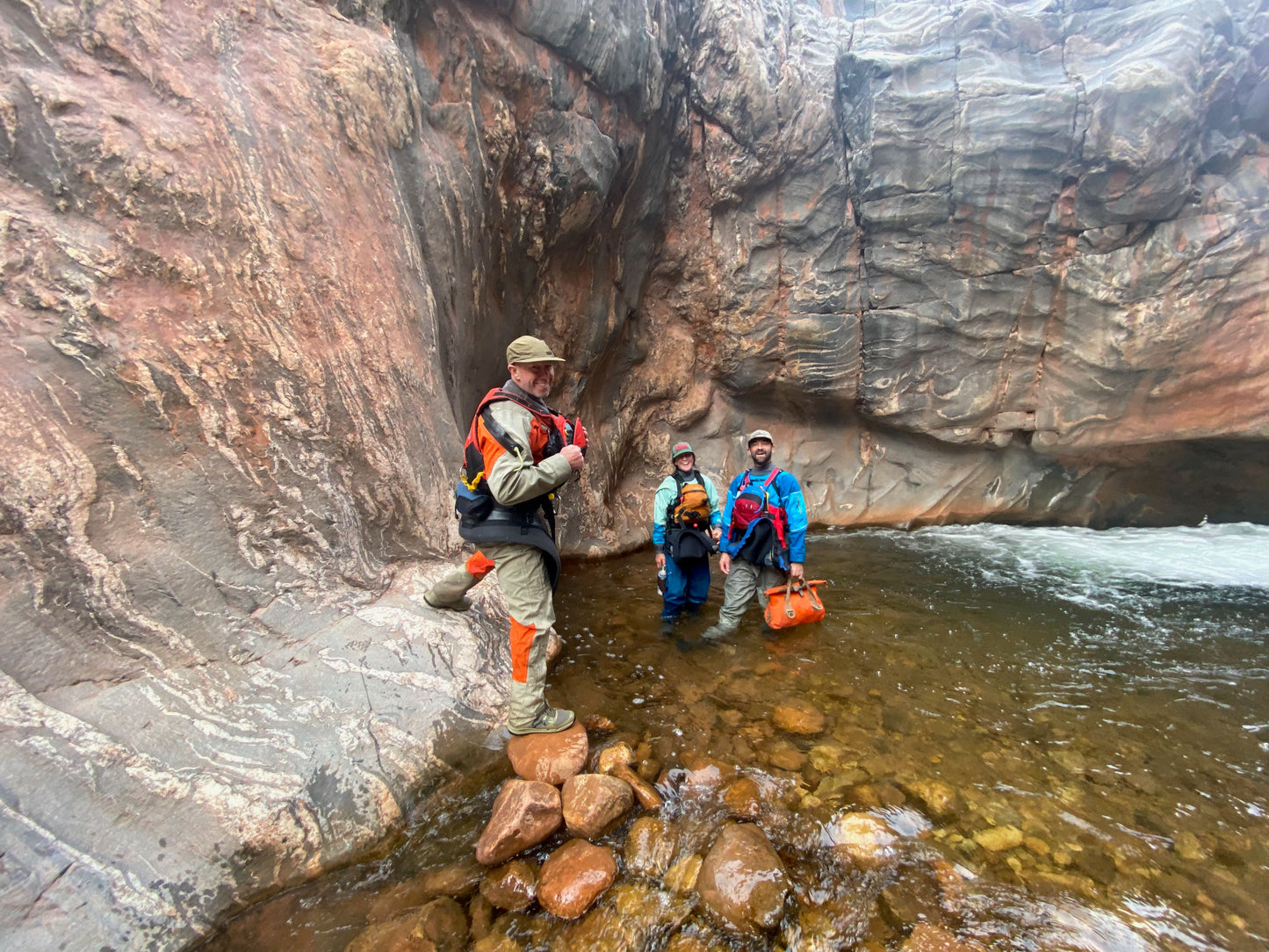 Three people in outdoor gear, including a 4CRS Grand Canyon Rental Drysuit, stand in shallow water near a canyon wall while one steps onto rocks in the foreground.