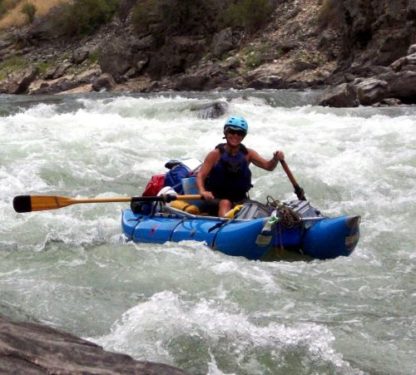 In a blue Cutthroat by Jacks Plastic Welding, a person skillfully navigates whitewater rapids with a paddle. Equipped with a helmet, they maneuver past rocky riverbanks showcasing the raft's surfing capabilities.