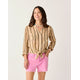 A woman wears the Carve Dylan Gauze Tunic, a tan and brown striped shirt tied at the waist, paired with pink shorts and layered necklaces, standing against a plain light background.