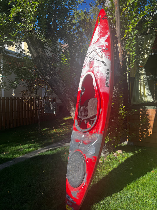 A Pyranha Fusion 10’ kayak from 4Corners Riversports stands upright on grass in a shaded backyard, perfect for self-supported trips, with trees and a wooden fence in the background.