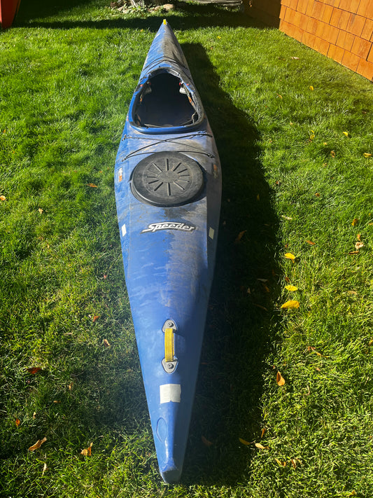A blue Pyranha Speeder 14' kayak from 4Corners Riversports with a covered storage hatch rests on green grass next to a wooden building.