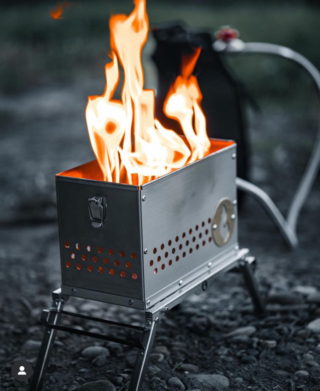 The LavaBox Hekla Portable Campfire / Stove Combo features bright flames on rocky ground, with a black bag and hose—ideal for outdoor cooking and perfect for your next adventure.