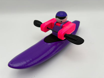 The Foamie Friends Foamie Creeker features a small toy figure in a purple kayak with a black paddle—wearing a purple cap and pink vest—perfect for river play and adventure.