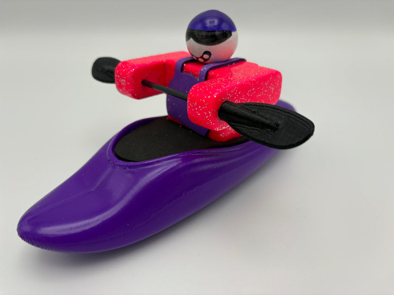A Foamie Friends toy figure in a helmet and life jacket steers the Foamie Play Boat, holding a black double-bladed paddle and ready for river adventures.