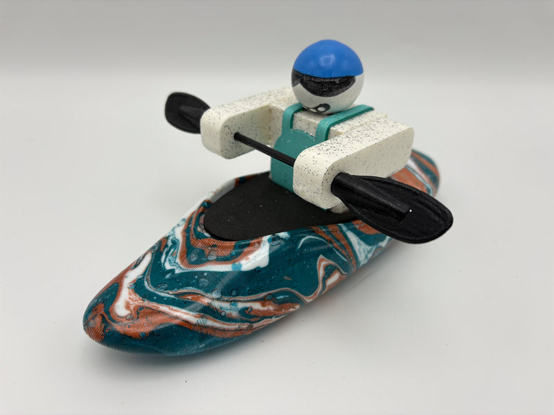 A Foamie Friends Swirl Play Foamie Boaters figure with a blue helmet and paddle sits in a marbled teal, white, and orange plastic play kayak against a plain background.