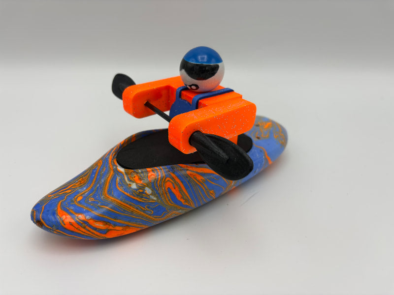 A Foamie Friends Swirl Play Foamie Boaters figure with a blue helmet and orange vest paddles in a blue and orange marbled plastic play boat against a plain light background.