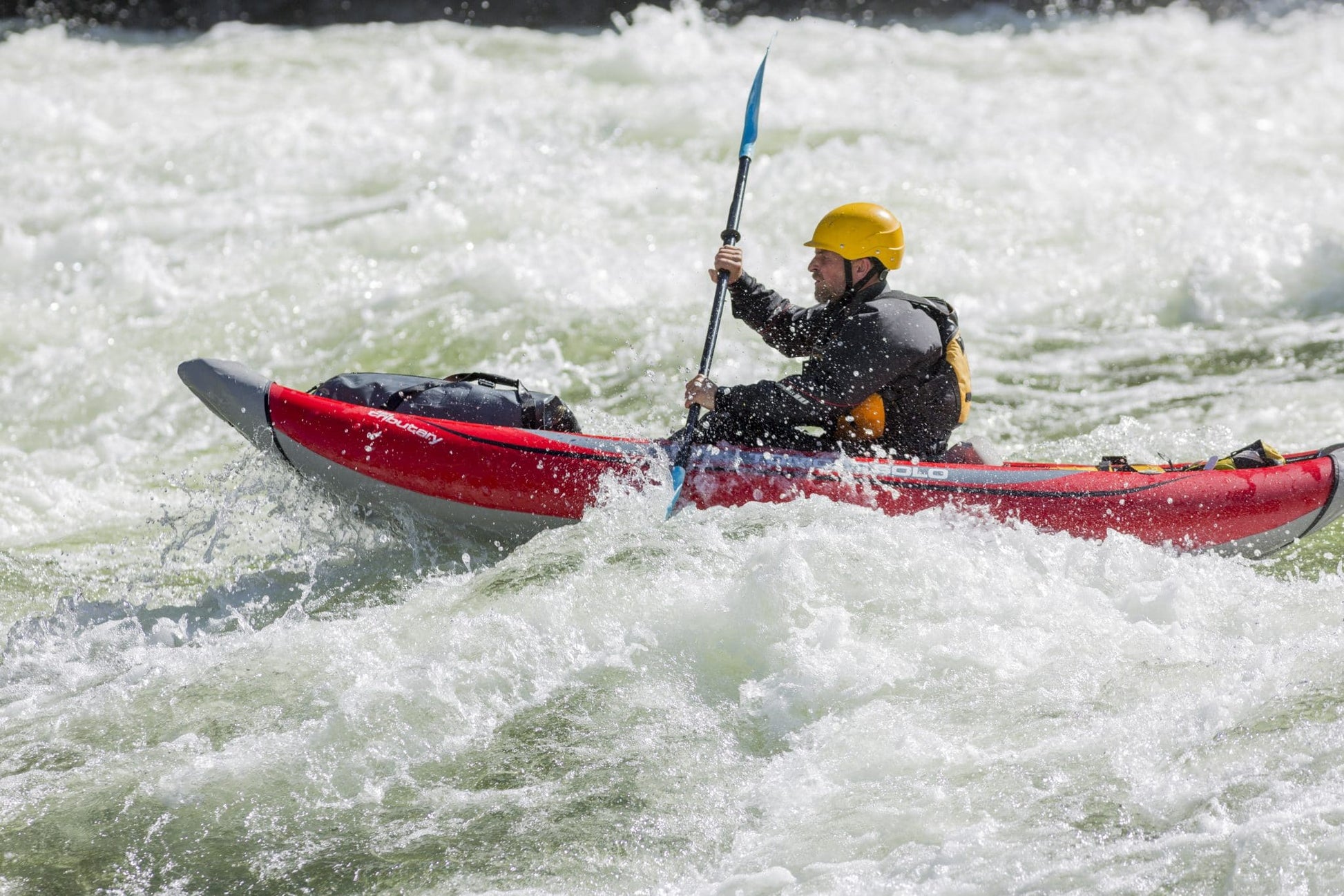 A person wearing a yellow helmet paddles a red 4CRS Rental Inflatable Kayak through whitewater rapids.