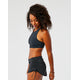A woman stands sideways, looking down and smiling, dressed in a black Carve Hoku Swim Skirt with adjustable hem and a black sports crop top, set against a plain white background.