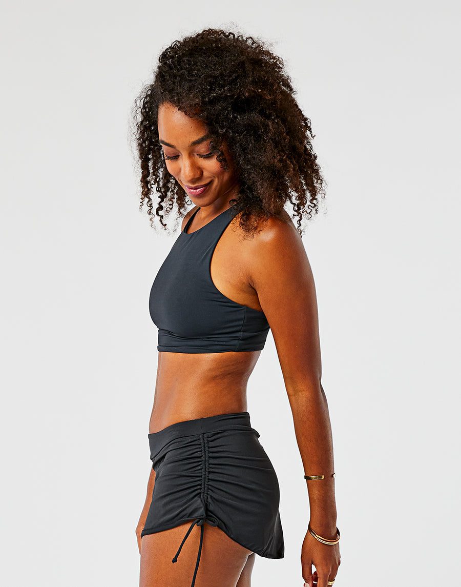 A woman stands sideways, looking down and smiling, dressed in a black Carve Hoku Swim Skirt with adjustable hem and a black sports crop top, set against a plain white background.