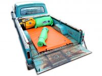 A vintage turquoise pickup truck bed contains a green sleeping bag, an orange Pickup Paco Pad by Jacks Plastic Welding, and two inflatable waterproof PVC pillows.