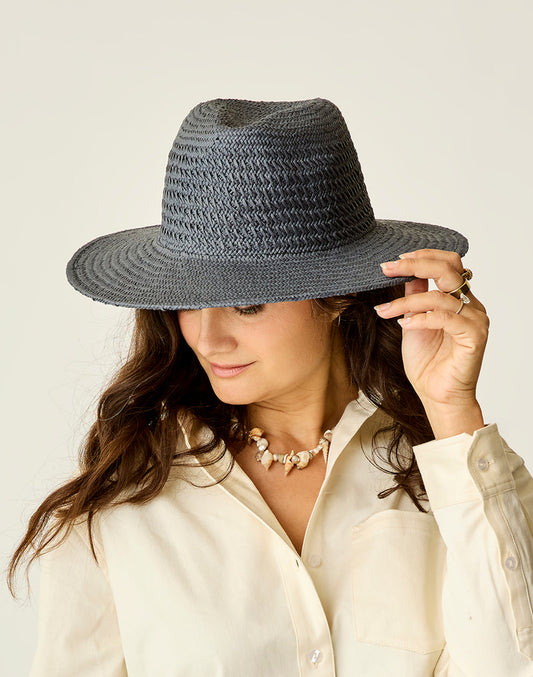 A woman in a cream blouse and shell necklace looks down while adjusting the Carve Soleil Hat, a wide-brimmed woven gray hat.