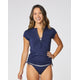 A woman smiles against a plain background wearing the Carve Dawson Rashguard, a navy quick-dry swim top with UPF 50+ sun protection and side ties, paired with a matching bikini bottom.