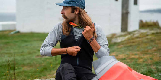 A person with long hair and a mustache sits outdoors, wearing a cap and zipping up a 4CRS Rental Paddle Gear wetsuit, with grass and a white building in the background.