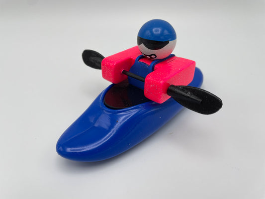 From the brand Foamie Friends, the Foamie Mini Playboat features a toy figure skillfully navigating river rapids, wearing a blue helmet and pink life vest. The figure is seated in a sleek blue kayak with an innovative hull design and black paddles, set against a clean white background.
