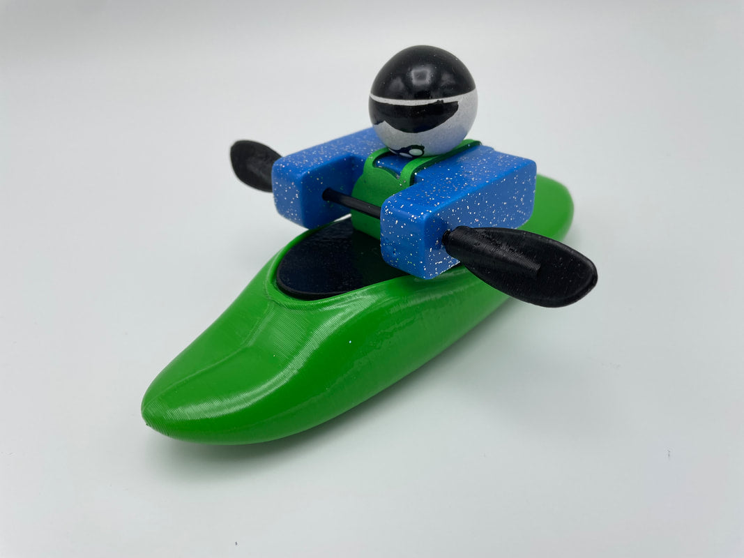 Foamie Friends Foamie Mini Playboat: A toy kayaker in a green boat with an innovative hull design, equipped with a black paddle, and wearing a blue jacket and black helmet against a white background.