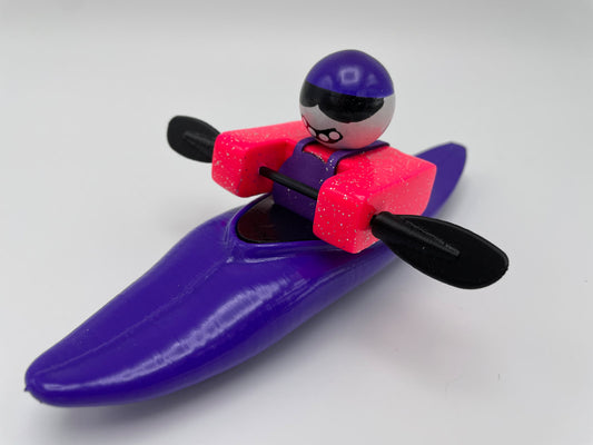 A Foamie Friends figurine, featuring a purple kayak that highlights the sleek hull design of the Foamie Mini Slice boats. Dressed in a pink vest and purple helmet, and donning black sunglasses while holding a paddle, this figurine brings together style and performance for an adventurous vibe.