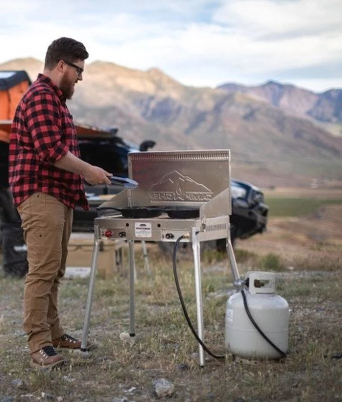 A man is cooking on a Camp Chef Mountaineer Camp Stove Leg Kit in front of a jeep.