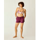 A woman stands barefoot against a plain light background wearing the Carve Bali Short in purple, paired with a striped bikini top, a beaded necklace, and bracelets.