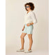 A woman wears a white knit sweater, Carve Oahu 6" Shorts in light blue stretch cotton corduroy, white loafers, sunglasses, and bracelets against an off-white background—the ideal foundation for an effortless vacation wardrobe.