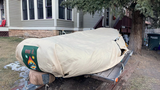 A boat sits on a trailer beside a house, protected by an Over It Raft Covers UV-stabilized cover, with part of the brand's green sticker reading “over it.” visible on the end.