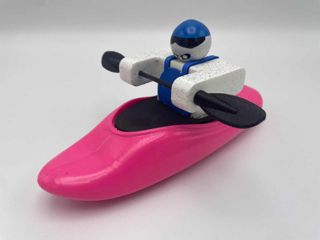 A toy figure depicting a kayaker paddling in a pink Foamie Play Boat, dressed in a blue and white outfit and using a black paddle, against a plain background. As part of the Foamie Friends collection, this piece embodies the excitement of river adventures for young explorers.