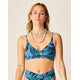 A woman wears the Carve Elba Top by Carve, a seamless bralette bikini with adjustable straps in a blue and green leaf pattern, paired with matching high-waisted bottoms and layered shell necklaces, standing against a plain background.