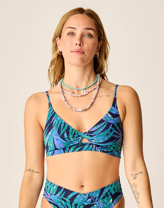 A woman wears the Carve Elba Top by Carve, a seamless bralette bikini with adjustable straps in a blue and green leaf pattern, paired with matching high-waisted bottoms and layered shell necklaces, standing against a plain background.
