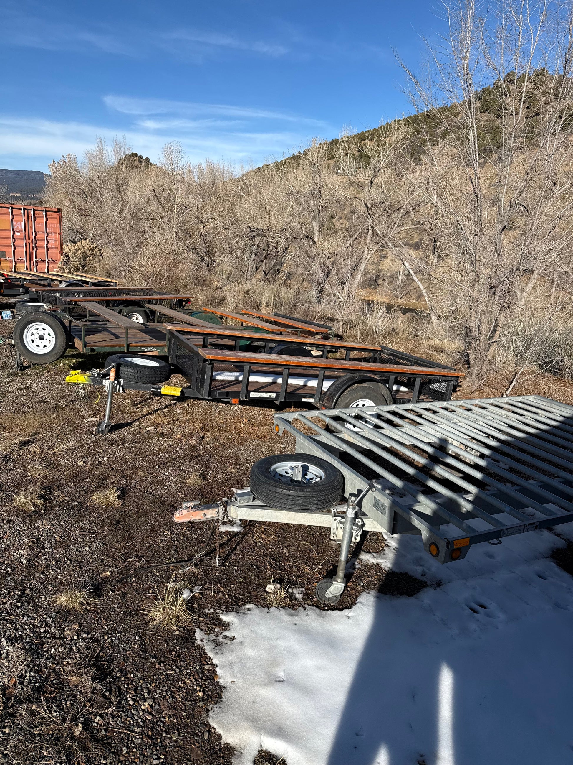 Two 4CRS Rental Trailer - Raft units, ideal for renting or hauling expedition rafts, sit on gravel and snow by bare trees. One trailer is tilted forward, casting a long shadow across the quiet winter setting.