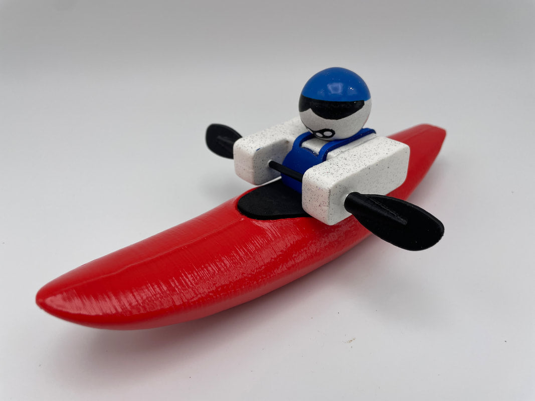 A Foamie Mini Creeker from the Foamie Friends brand is set for river adventures, seated in a red kayak with a blue helmet and black paddles against a simple backdrop.