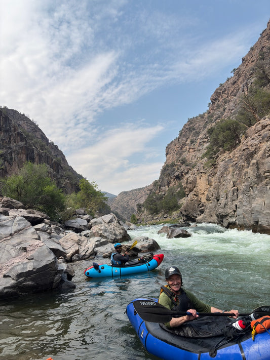 Two people in 4CRS Rental Packrafts paddle down a rocky river canyon under a partly cloudy sky, surrounded by steep rock walls and sparse vegetation.