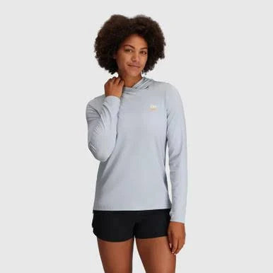 A woman wears the Outdoor Research ActiveIce Spectrum Sun Hoodie in light gray, paired with black shorts, standing against a plain light background.